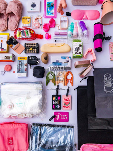 A flat-lay collection of various travel accessories and toiletries on a white background, including a pink sleep mask, fluffy slippers, a yellow clutch, toiletry bottles, a pink travel towel, and other packing essentials.